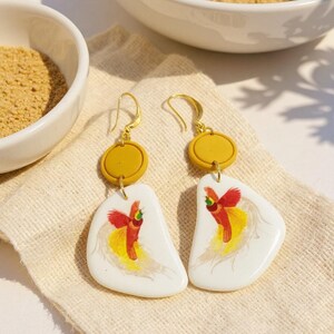 May include: A pair of gold-tone earrings. Each earring features a mustard-yellow disc and a white, irregular-shaped pendant with a red and yellow bird design. The earrings rest on a beige cloth, with bowls of brown powder in the background.