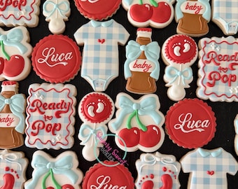 May include: A collection of decorated sugar cookies featuring baby-themed designs including red and white rattles with footprints, red circles with the name Luca, blue and white gingham onesies, red cherries with blue bows, and soda bottles.