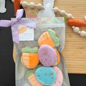 May include: A clear cellophane bag tied with a lavender ribbon contains Easter-themed cookies. The cookies are shaped like carrots and speckled eggs in orange, purple, blue, and pink. A tag with "Happy Easter" is attached.