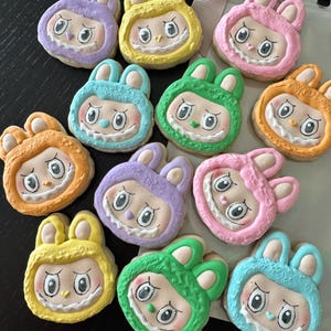 May include: A collection of colorful, bunny-shaped cookies. Each cookie features a cartoon face with large eyes and a wide grin, and is decorated with pastel frosting in shades of yellow, pink, green, blue, orange, and purple.