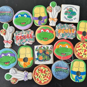 May include: A collection of decorated sugar cookies featuring Teenage Mutant Ninja Turtles characters and pizza slices. The cookies are round, square, and turtle shell-shaped, with green, yellow, and red icing.