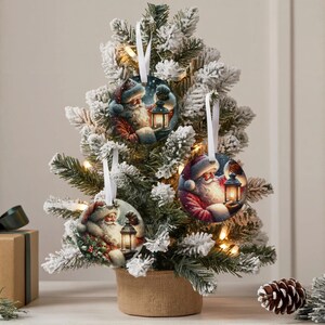Puede incluir: Tres adornos redondos de cer&aacute;mica con ilustraciones de Santa Claus sosteniendo una linterna brillante. Cada adorno est&aacute; suspendido por una cinta blanca y cuelga de las ramas de un peque&ntilde;o &aacute;rbol de Navidad artificial nevado.