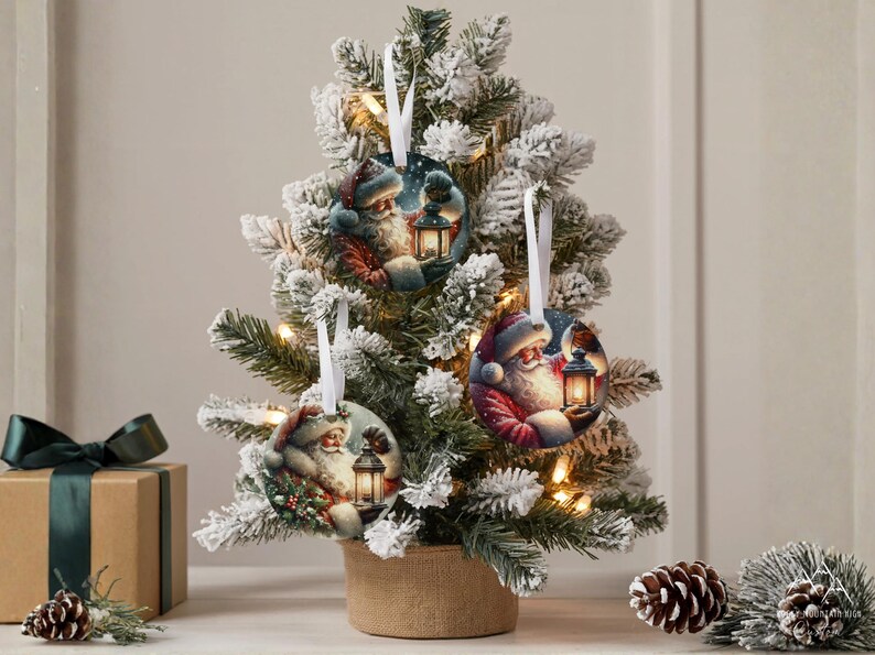 Puede incluir: Tres adornos redondos de cer&aacute;mica con ilustraciones de Santa Claus sosteniendo una linterna brillante. Cada adorno est&aacute; suspendido por una cinta blanca y cuelga de las ramas de un peque&ntilde;o &aacute;rbol de Navidad artificial nevado.