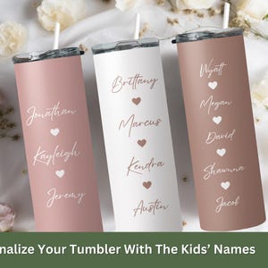 May include: Three personalized stainless steel tumblers with pink, white, and brown finishes. Each tumbler features a name and heart design. The tumblers are arranged in a row on a white surface with flowers in the background. The text on the tumblers reads: "Brittany, Marcus, Kendra, Austin", "Jonathan, Kayleigh, Jeremy", and "Wyatt, Megan, David, Shawna, Jacob".