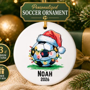 May include: A round white ceramic ornament featuring a soccer ball wearing a red Santa hat and wrapped in colorful Christmas lights. The name NOAH and the year 2026 are printed in black text below the ball. A gold string is attached at the top.