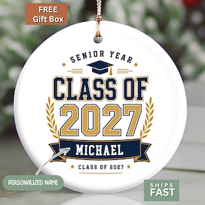 May include: A round white ceramic ornament featuring a navy blue graduation cap icon, the text SENIOR YEAR, CLASS OF 2027, and the name MICHAEL printed in navy blue and gold. The design includes gold laurel branches and a navy blue banner.
