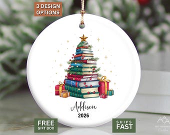 May include: A round white ceramic Christmas ornament featuring a stack of colorful books arranged in the shape of a tree, topped with a gold star. The design includes small gift boxes at the base, the name Addison, and the year 2026 in black text.