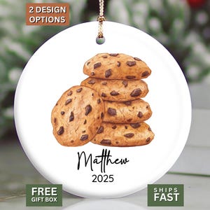May include: A round white ceramic ornament featuring a watercolor illustration of a stack of four chocolate chip cookies. Below the cookies, the name Matthew is printed in black script, followed by the year 2025 in a simple black font.