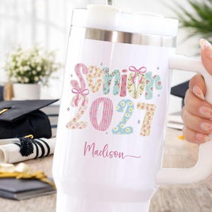 May include: A white insulated tumbler with a handle featuring the text Senior 2027 in a floral and striped pattern with pink bows. The name Madison is printed in pink script below the text. The tumbler has a clear lid and a metal straw.