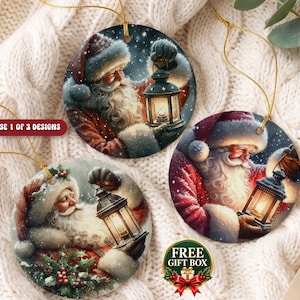 Puede incluir: Tres adornos navide&ntilde;os redondos de cer&aacute;mica con ilustraciones de Santa Claus sosteniendo una linterna brillante. Cada adorno tiene un cord&oacute;n dorado. El texto CHOOSE 1 OF 3 DESIGNS y FREE GIFT BOX es visible en la imagen.