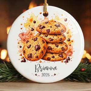 May include: A round white ceramic ornament featuring a watercolor illustration of a stack of four chocolate chip cookies. The name Brianna and the year 2026 are printed in black script and sans-serif font below the cookies. A gold string is attached.