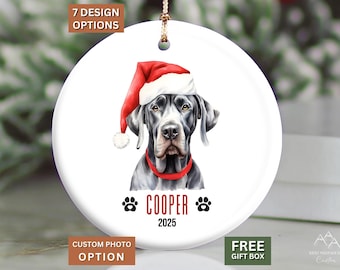 May include: A round white ceramic ornament featuring a watercolor illustration of a black Great Dane wearing a red Santa hat and a red collar. The text COOPER is printed in red below the dog, flanked by two black paw prints, with 2025 below.