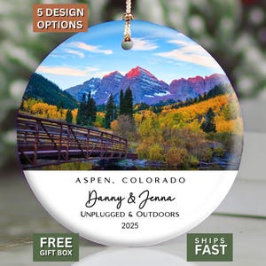 May include: A round ceramic ornament featuring a landscape photograph of the Maroon Bells in Aspen, Colorado, with a wooden bridge and autumn trees. The white lower section displays the text: ASPEN, COLORADO, Danny & Jenna, UNPLUGGED & OUTDOORS, 2025.