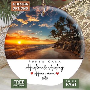 May include: A round ceramic ornament featuring a sunset beach scene with palm trees and ocean waves. The white lower section displays the text "PUNTA CANA", "Hudson & Audrey", "Honeymoon" with two red hearts, and the year "2025" in black lettering.