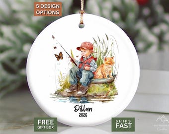 May include: A round white ceramic ornament featuring a watercolor illustration of a child in a red hat and vest fishing with a cat nearby. The design includes butterflies, tall grass, and the printed text "Dillan" and "2026" at the bottom.