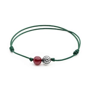 Könnte beinhalten: Ein grünes Armband aus Schnur mit einer roten Edelsteinperle und einem silbernen Anhänger mit Blumenmuster.