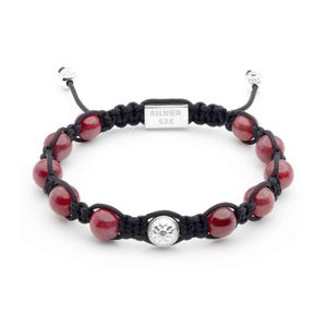 Könnte beinhalten: Ein Armband aus roten Edelsteinen mit einem silbernen Anhänger und einer schwarzen Schnur. Der Anhänger hat ein Blumenmuster und die Schnur ist verknotet. Das Armband hat ein silbernes Schild mit der Aufschrift "SILVER $25".