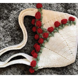 Op de afbeelding: Een witte gehaakte handtas met een decoratieve rank van kleine rode rozen en groene blaadjes langs de zijkant en het handvat. De tas is gemaakt met een gestructureerd steekpatroon en bevat twee bijpassende witte gehaakte schouderbanden.