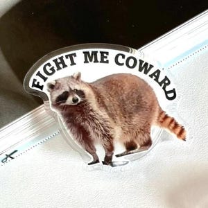 May include: A white sticker with a raccoon image and the text "Fight Me Coward" in black lettering.