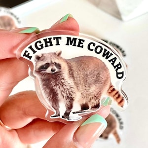May include: A clear sticker with a raccoon illustration and the text "Fight Me Coward" in black lettering.