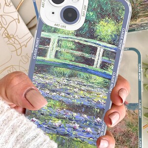 May include: Clear phone case featuring a painting by Claude Monet. The case showcases a bridge over water with lily pads, in shades of green, blue, and white. The case has the words "ART LIFE" and "CLAUDE MONET" printed on it.