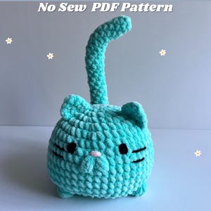 Pode incluir: Um brinquedo de pelúcia de gato azul turquesa crochetado com olhos pretos e um nariz branco. O gato tem uma cauda longa e está sentado em uma superfície branca. Padrão PDF sem costura.