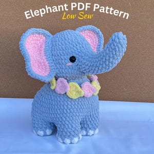 May include: A crocheted gray elephant toy with pink ears and a yellow and pink flower garland around its neck. The toy is sitting on a white surface. Elephant PDF Pattern Low Sew