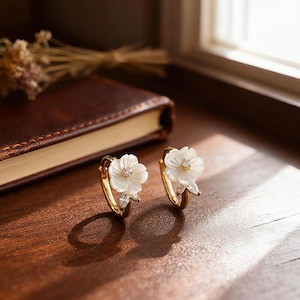 Könnte beinhalten: Ein Paar goldene Creolen, jeweils verziert mit einem weißen Blumenmuster und einem zentralen Edelstein. Die Ohrringe sind auf einer warmen Holzoberfläche platziert, mit einem Buch und getrockneten Blumen im Hintergrund. Ein zartes Accessoire.