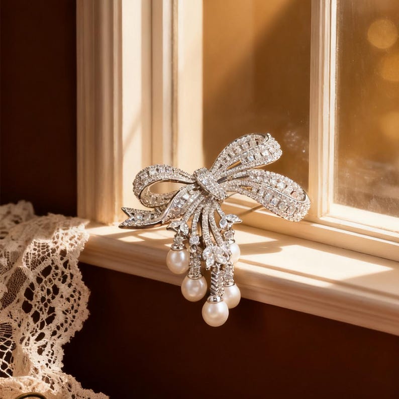 Puede incluir: Broche plateado con forma de lazo, incrustado con cristales brillantes. El dise&ntilde;o del lazo presenta hilos colgantes de perlas, creando un accesorio elegante. El broche se exhibe en un alf&eacute;izar.