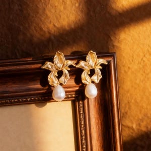 Op de afbeelding: Een paar goudkleurige oorbellen in bloemvorm met een druppelvormige, witte parel. De oorbellen worden gepresenteerd op een houten lijst tegen een warme, gouden achtergrond. De oorbellen hebben een gedetailleerd, getextureerd ontwerp.