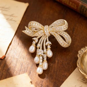 Puede incluir: Un broche dorado en forma de lazo, incrustado con cristales brillantes. El dise&ntilde;o del lazo presenta perlas colgantes en forma de l&aacute;grima. El broche est&aacute; sobre una superficie de madera oscura, con un libro y papel vintage en el fondo.