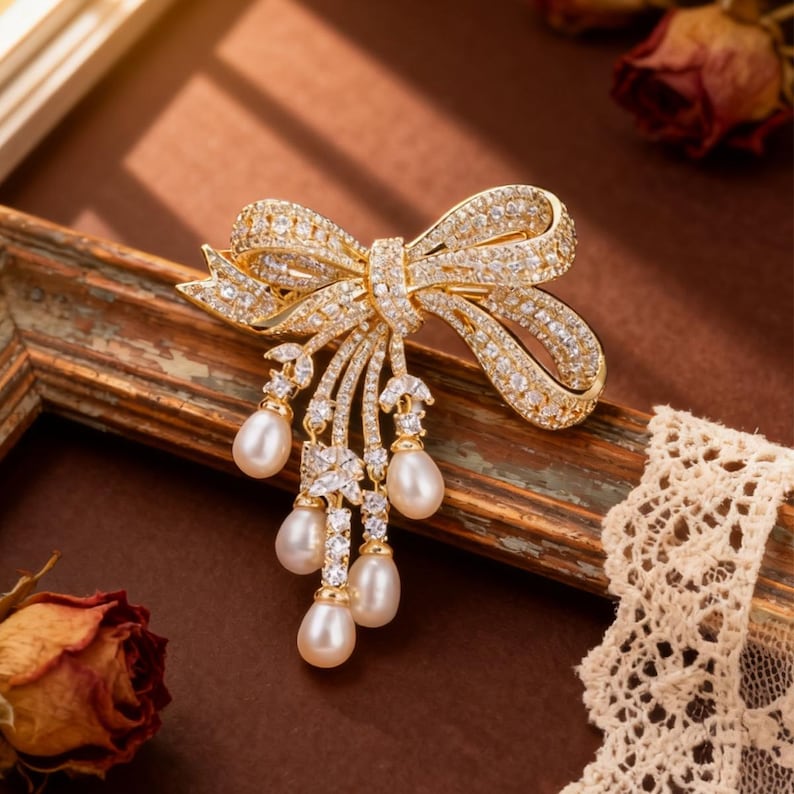 Puede incluir: Broche dorado con forma de lazo adornado con cristales brillantes y perlas de imitaci&oacute;n colgantes. El dise&ntilde;o del lazo est&aacute; detallado con intrincados engastes de cristal, y el broche presenta cinco gotas de perlas en forma de pera. Joya elegante para ocasiones especiales.