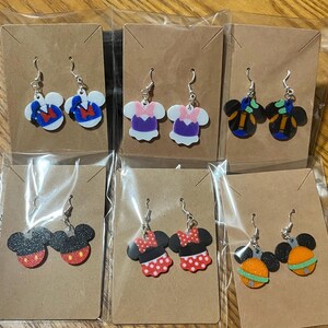 May include: A collection of six pairs of dangle earrings, each featuring a different cartoon character design. The earrings are in clear plastic packaging and attached to brown cardstock. Designs include Mickey Mouse, Minnie Mouse, and other character-themed earrings.
