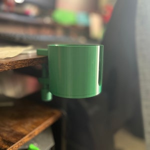 May include: A green plastic cup holder mounted on a desk. The holder is designed to hold a cup or mug.