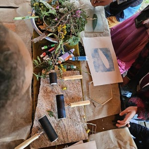 Op de afbeelding: Een tafel voor botanisch printen met gereedschap en materialen. Inktrollers, verftubes en geperste bladeren en bloemen zijn gerangschikt. Er zijn verschillende afdrukken zichtbaar, met blad- en veerafdrukken.