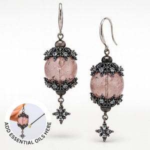 May include: A pair of dangle earrings with a pink, orb-shaped center. The earrings have a dark metal filigree design with small, clear stones. The text "ADD ESSENTIAL OILS HERE" is in the lower left corner.
