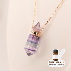 May include: A purple and green fluorite essential oil bottle pendant with a gold chain. The bottle has a pointed top and a gold-colored metal band. The image includes a small bottle with the text "FREE SAMPLE Essential Oil Blend".