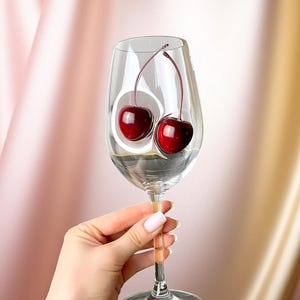 Pode incluir: Um copo de vinho transparente com duas cerejas maduras dentro, segurado por uma mão. As cerejas são de cor vermelha escura, e o copo está sobre um fundo de tecido rosa e dourado.