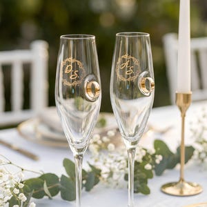 Peut inclure: Deux flûtes à champagne transparentes avec des porte-anneaux dorés. Chaque verre porte un monogramme doré et une bague dorée. Une bougie blanche dans un support doré est posée sur une nappe blanche, avec de la verdure et des fleurs.