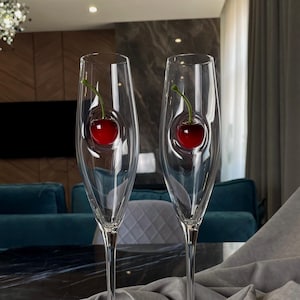 Puede incluir: Dos copas de champán transparentes con un diseño de cereza roja en el interior, sobre una bandeja de espejo. Las copas tienen tallos largos y están sobre tela gris. El fondo muestra un salón moderno.