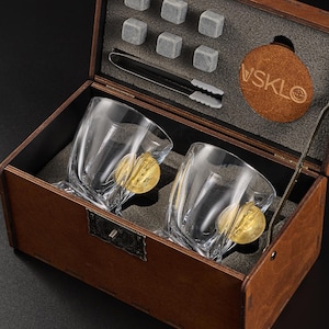 Peut inclure: Un coffret en bois contenant deux verres à whisky transparents avec des accents dorés, six glaçons en pierre grise et une pince. La boîte comprend également un dessous de verre rond en cuir avec le texte "VSKLO".