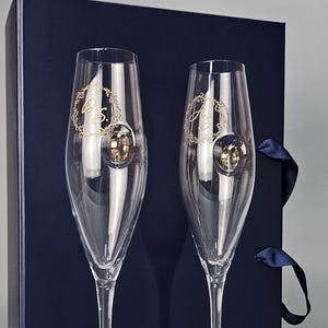 Peut inclure: Deux flûtes à champagne transparentes avec des initiales dorées et un motif de couronne. Elles sont présentées devant un coffret cadeau bleu marine avec un ruban assorti. Les verres sont élégants et adaptés à une occasion spéciale.