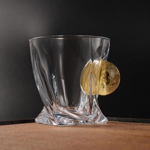 May include: A clear, sculpted glass with a gold Bitcoin coin attached. The glass has a square base and a flared, wavy design. The Bitcoin coin is prominently displayed on the side of the glass, against a dark background.
