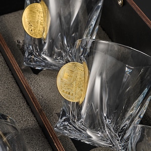 May include: Set of clear, faceted whiskey glasses with a gold-colored Bitcoin coin attached. The glasses are displayed inside a dark gray lined box, suggesting a gift set. The glasses have a unique, angular design.