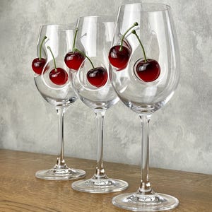 Può includere: Tre bicchieri da vino trasparenti con steli lunghi, ciascuno con una singola ciliegia rossa dall'aspetto realistico. Le ciliegie hanno gambi verdi e sono posizionate all'interno delle coppe. I bicchieri sono disposti su una superficie di legno.