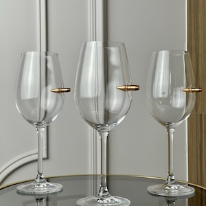 Puede incluir: Tres copas de vino transparentes con diseño de bala. Las copas están sobre una mesa redonda con tapa de cristal y marco de metal dorado. El diseño de la bala es de color cobre.