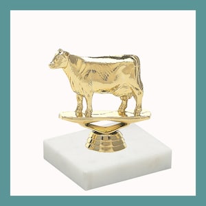 May include: Gold cow trophy on a white marble base. The cow is standing on a gold platform.