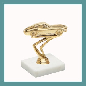 May include: Gold-colored trophy featuring a stylized Corvette car on a swirling base. The base is mounted on a white marble square.