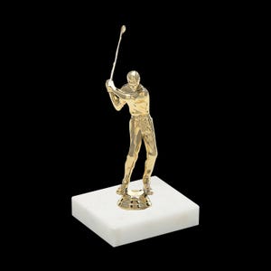May include: A gold-colored golf trophy featuring a golfer swinging a club. The trophy sits on a white marble base.