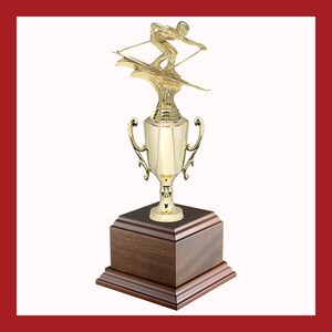 May include: A gold-colored trophy featuring a skier in action atop a cup-shaped body with handles. The trophy sits on a dark brown wooden base, ideal for sports awards and achievements.